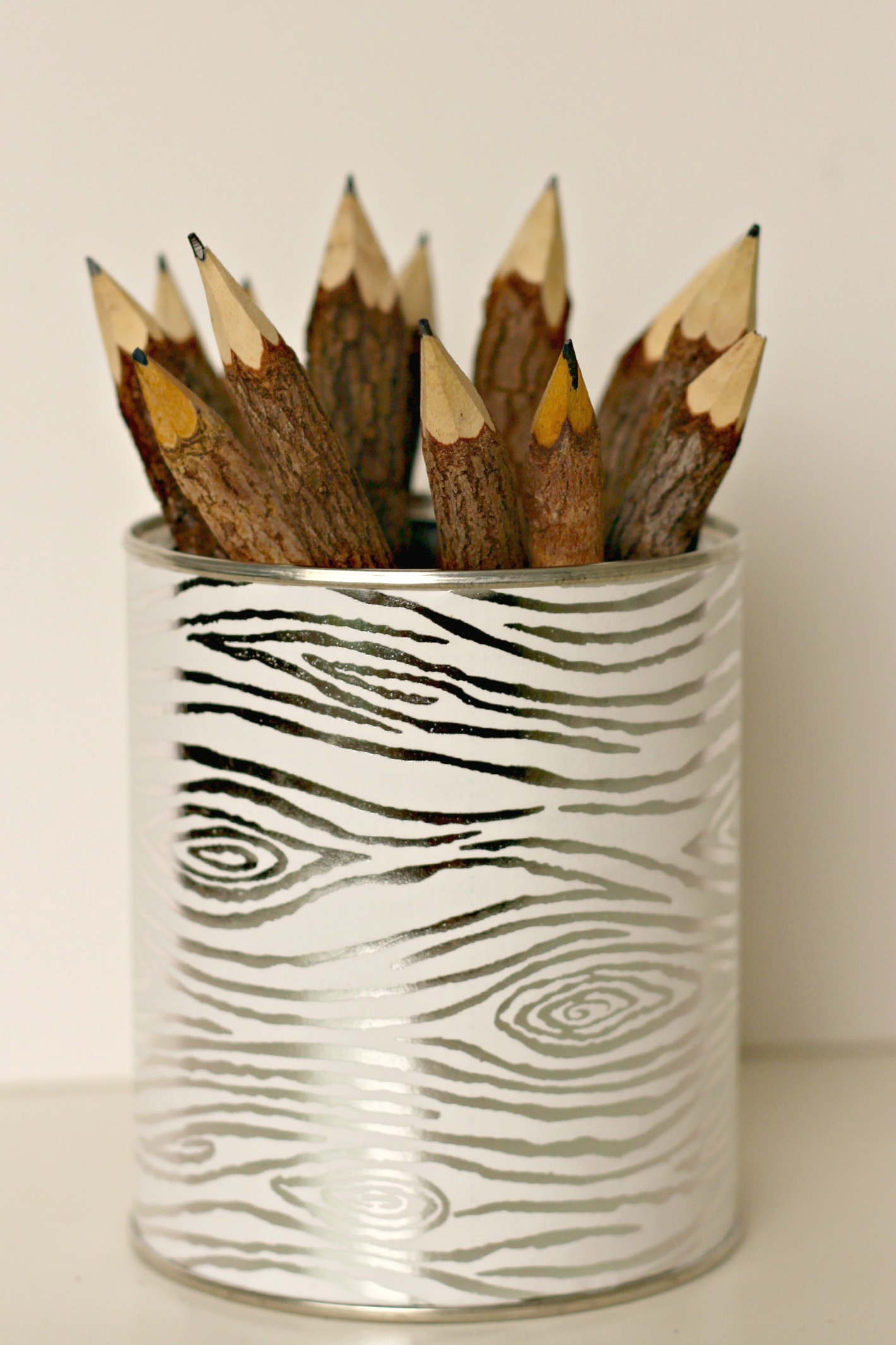 Tall tin can wrapped in silver and white wood grain paper holding a bundle of rustic twig pencils on a white surface